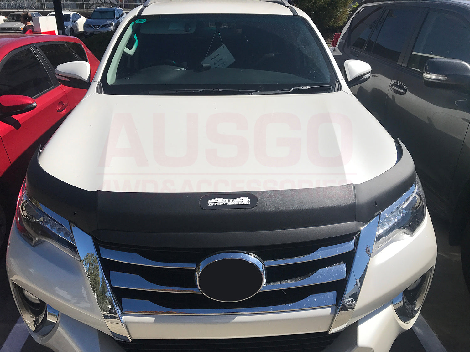 Front view of a white 4x4 SUV fitted with an Ausgo bonnet protector, parked between vehicles in a sunlit outdoor lot.