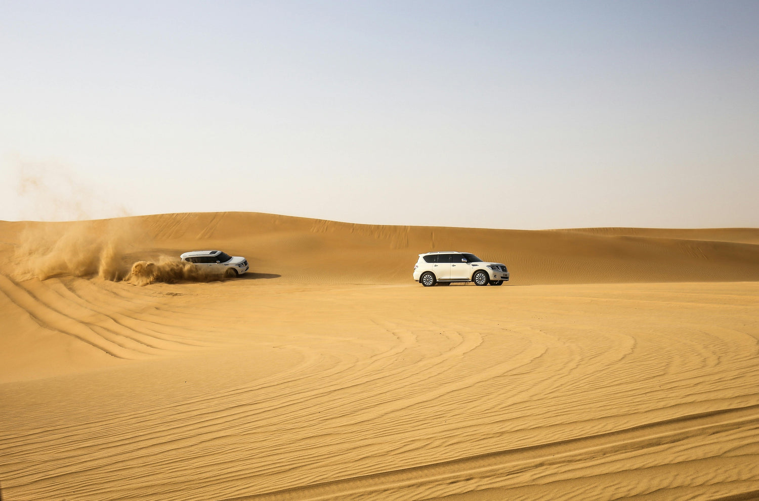Two white 4x4 vehicles driving through sandy desert dunes, one kicking up sand while off-roading under a clear blue sky.
